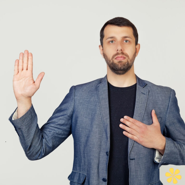 Man in a suit raising his hand with a serious expression, other hand on his chest.