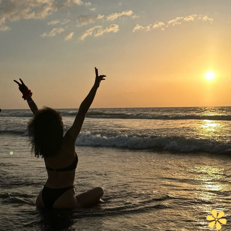 Person with curly hair sits in water at sunset, arms raised in joy.
