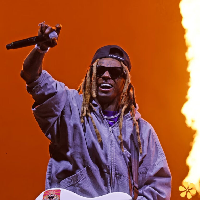 A performer with long dreadlocks in a grey hoodie, smiling and holding a microphone with flames behind.