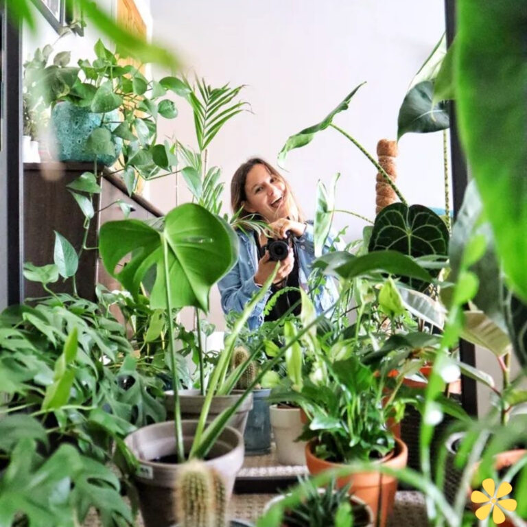 Woman smiling amidst lush indoor plants, holding a camera, surrounded by greenery.