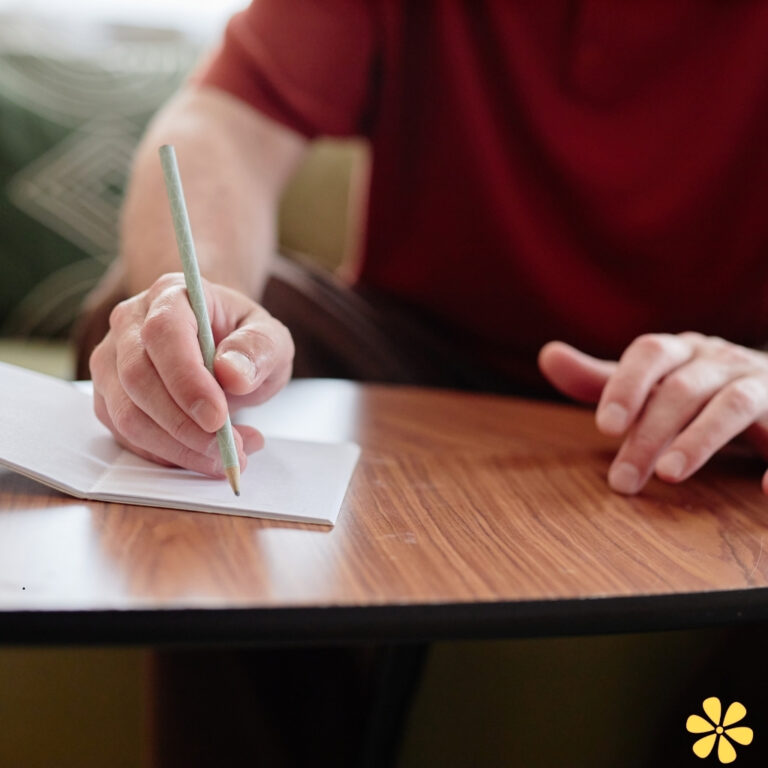 A hand grips a green pencil, poised over a blank notebook on a wooden table.
