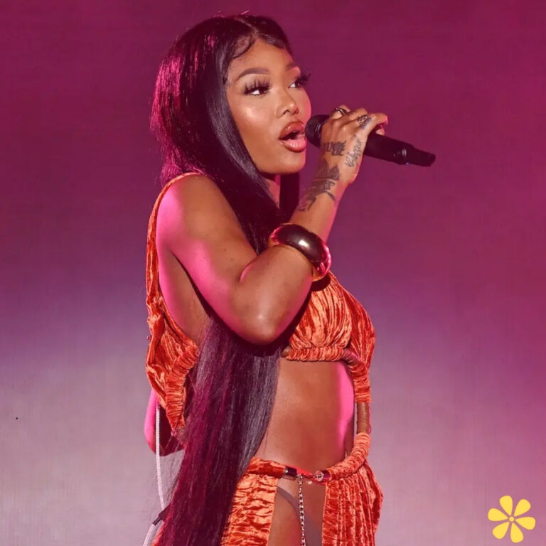 A performer with long black hair sings passionately into a microphone, dressed in a striking orange outfit.