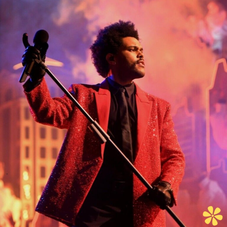 A performer in a dazzling red sparkly suit stands confidently with a microphone, dramatic lighting surrounding him.