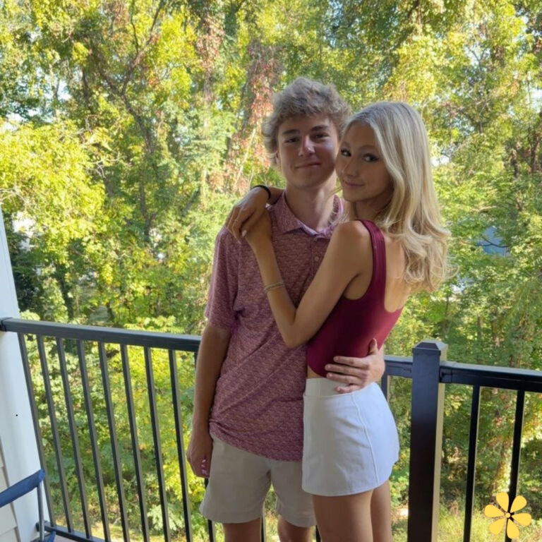 A young couple embraces on a balcony surrounded by lush greenery, smiling softly at the camera.