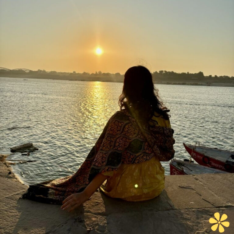Woman sitting by the water, watching the sunset with a colorful scarf flowing.