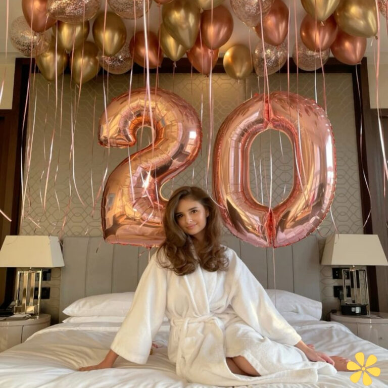 Young woman in a robe sits on a bed, surrounded by large rose gold balloons that say '20'.