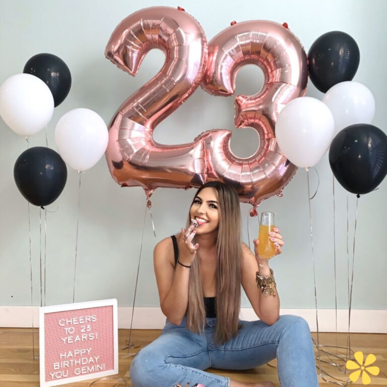 A young woman sits with a playful smile, holding a drink, surrounded by large balloons spelling '23'.