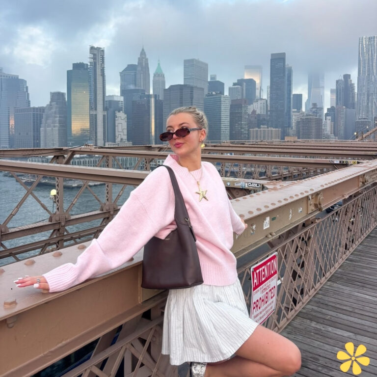 Woman in a pink sweater and white skirt poses confidently on a bridge with a city skyline behind her.