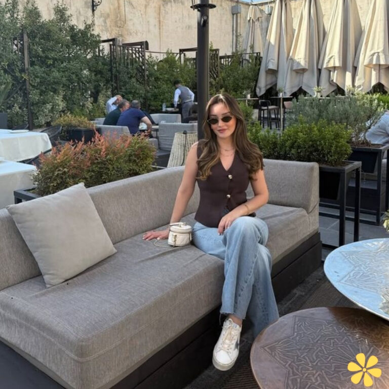 A woman in a stylish outfit sits comfortably on a gray lounge sofa at an outdoor café, sunglasses on.