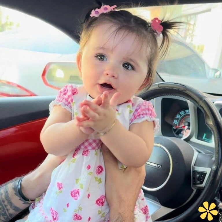 A joyful baby claps her hands while sitting in a car, adorned with a dress decorated with roses.