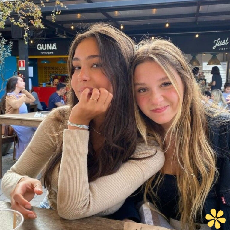 Two friends smiling together at a café, one resting chin on hand, the other with long hair.