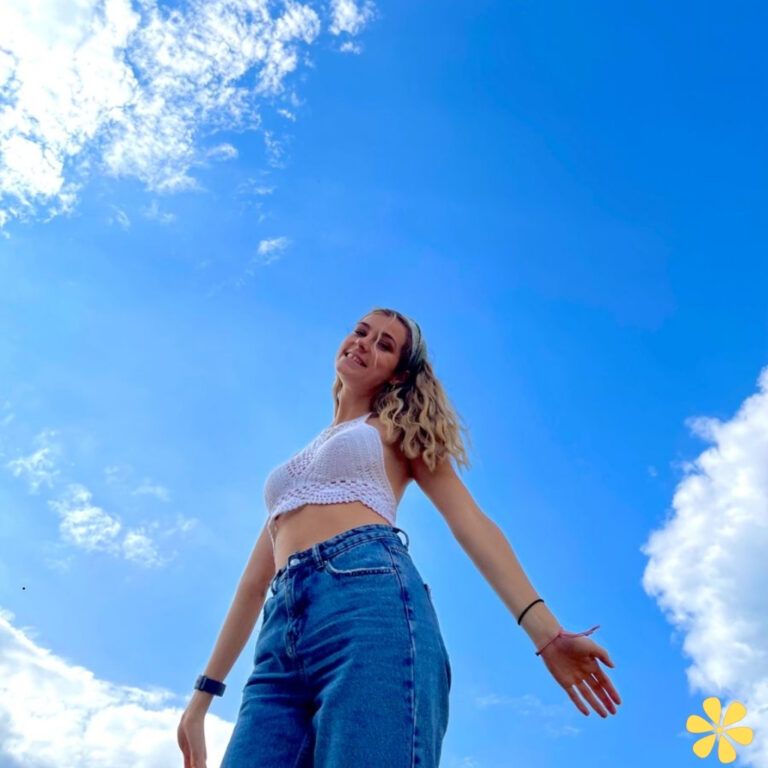 A person in a crochet top and high-waisted jeans stands against a bright blue sky with fluffy clouds.