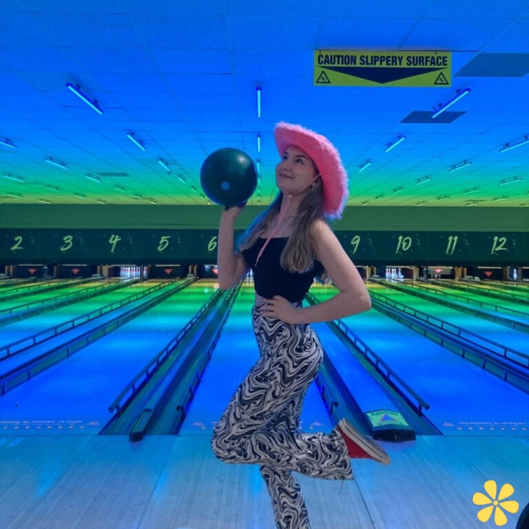 Woman in a pink hat holds a bowling ball, striking a playful pose in a brightly lit bowling alley.