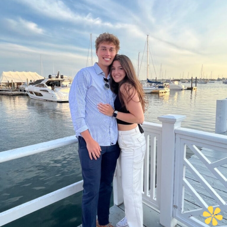 Young couple smiles together at a marina, boats in the background under a beautiful sky.