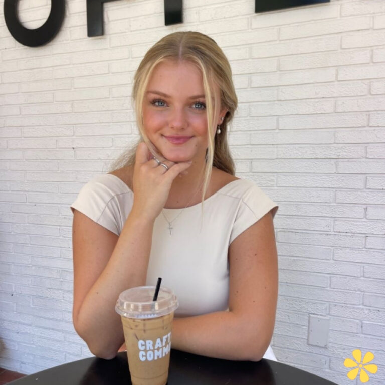 Smiling young woman with a drink, hand on chin, sitting at a table against a white wall.
