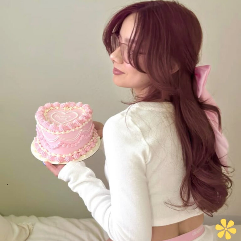 A woman with long brown hair holds a pink heart-themed cake, wearing a white top with a pink bow in her hair.