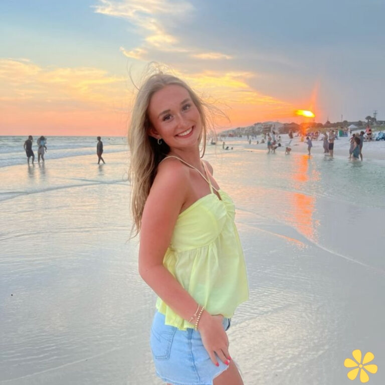 A young woman in a yellow top smiles at the camera while standing on a beach at sunset.