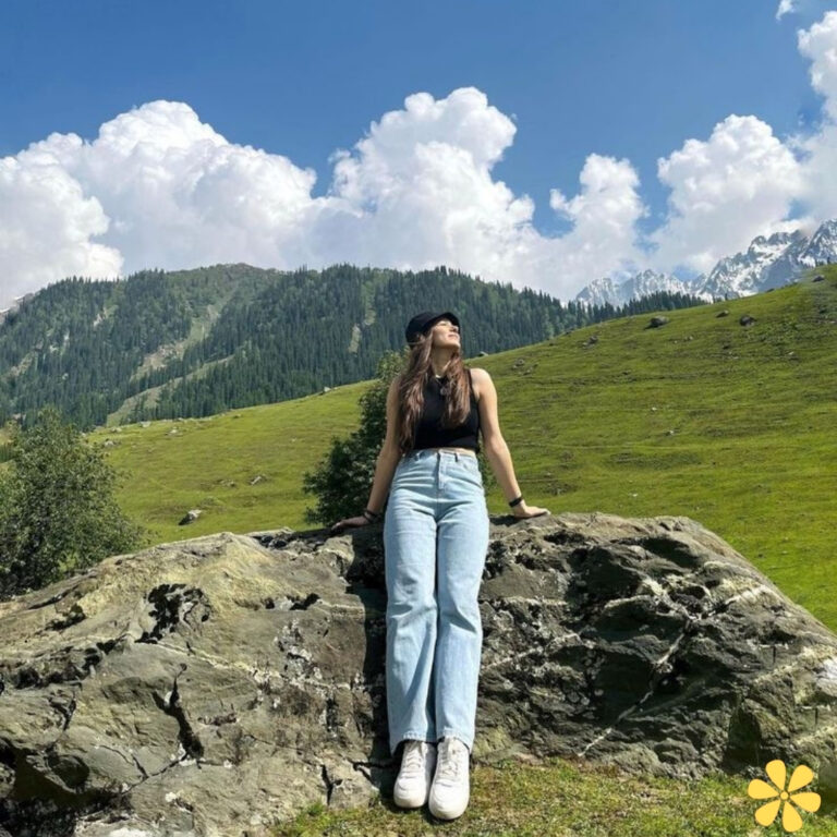 Young woman in a black tank top and light jeans stands on a rock, surrounded by lush greenery and mountains.