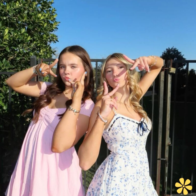 Two friends posing playfully outdoors, one in a pink dress, the other in a floral dress, both making peace signs.
