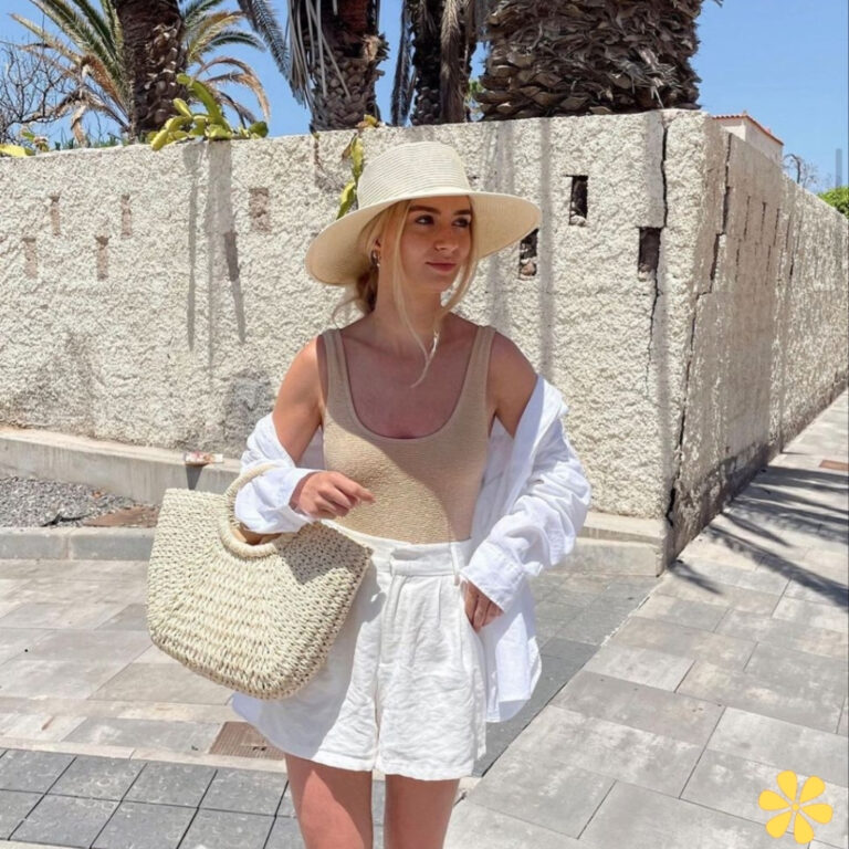 Woman in a sun hat and beige top, holding a woven bag against a textured wall.
