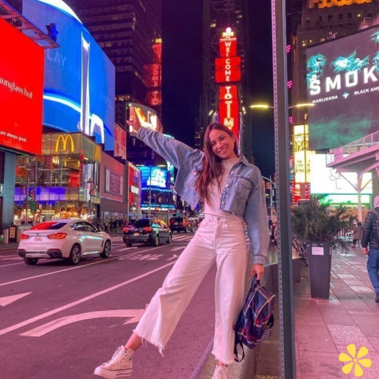 A young woman stands joyfully in Times Square, wearing a denim jacket and white pants, posing with one arm raised.