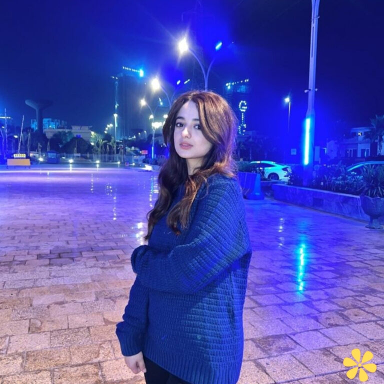 Young woman in a cozy blue sweater stands confidently at night, illuminated by soft blue lights.