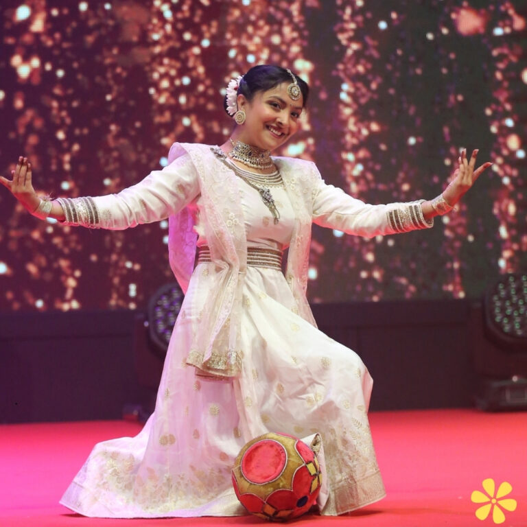 A woman in a white traditional outfit joyfully poses on stage with a soccer ball, surrounded by sparkling lights.