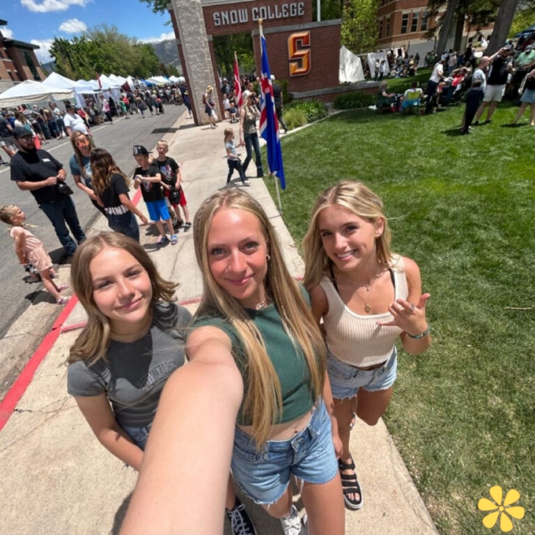 Three friends smiling at a college event, surrounded by people and tents in the background.
