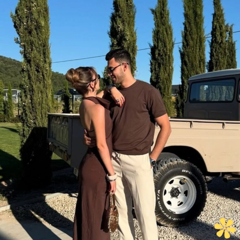 Couple smiling in casual outfits, standing by a vintage truck and tall cypress trees.