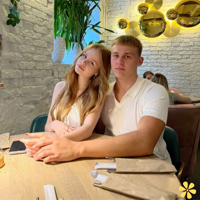 A couple sits closely together at a restaurant table, smiling softly, with greenery in the background.