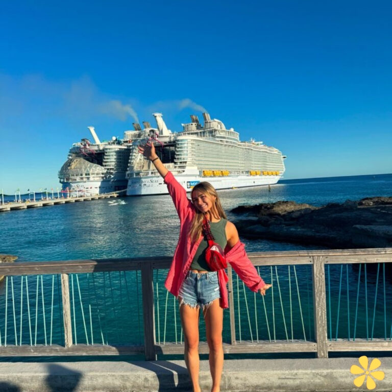 Girl with a red jacket joyfully poses by the ocean, a large cruise ship behind her.