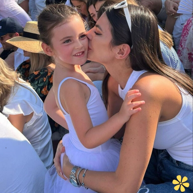 A woman kisses a smiling girl in a white dress, both look joyful in a crowd.