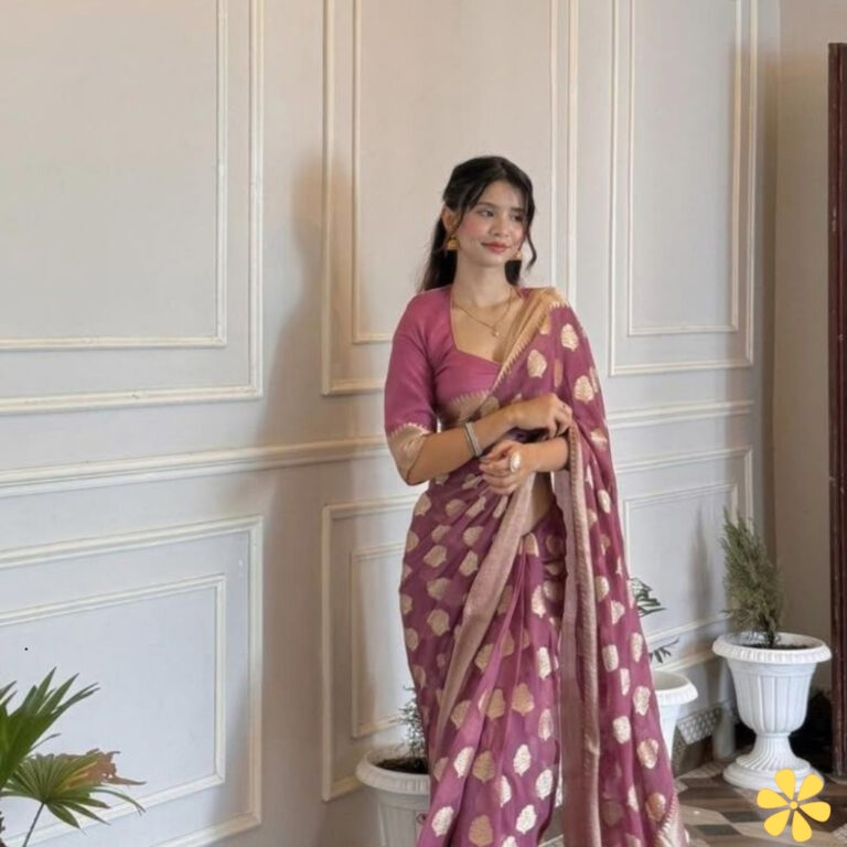 Young woman in a pink saree stands gracefully in a bright room with decorative plants.
