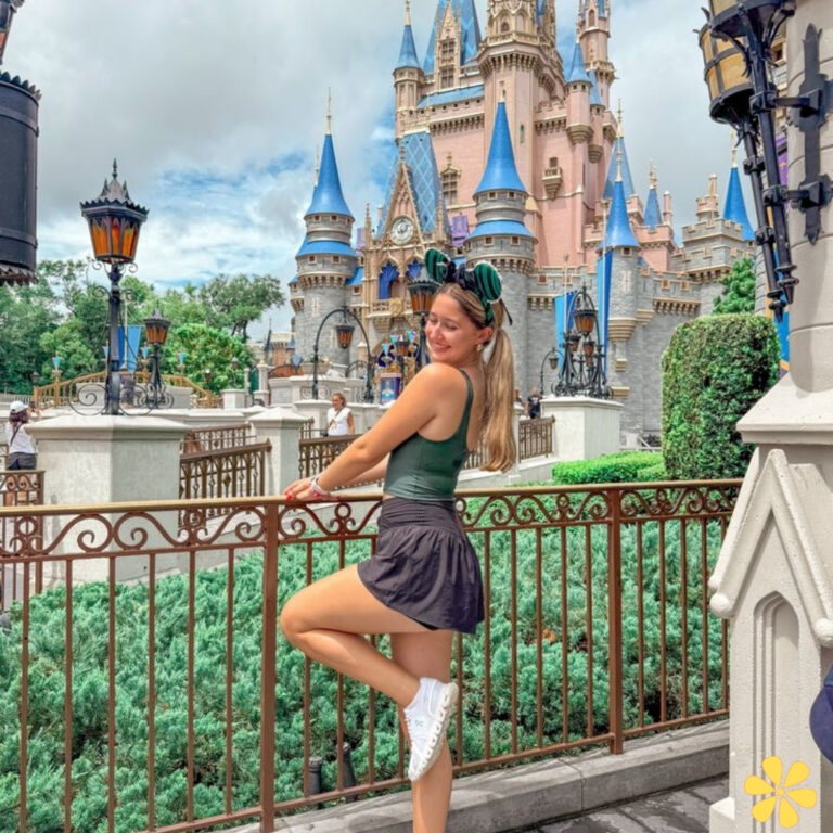 A young woman poses playfully in front of a fairytale castle, wearing a green top and a black skirt.