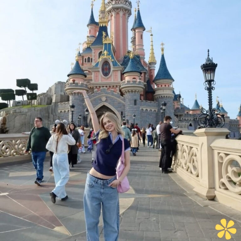 Smiling person in a blue sweater and jeans poses with one arm raised in front of a castle.