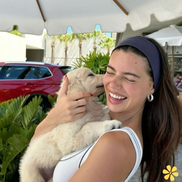 Woman joyfully holds a puppy, both smiling. Sunlight filters through trees behind them.