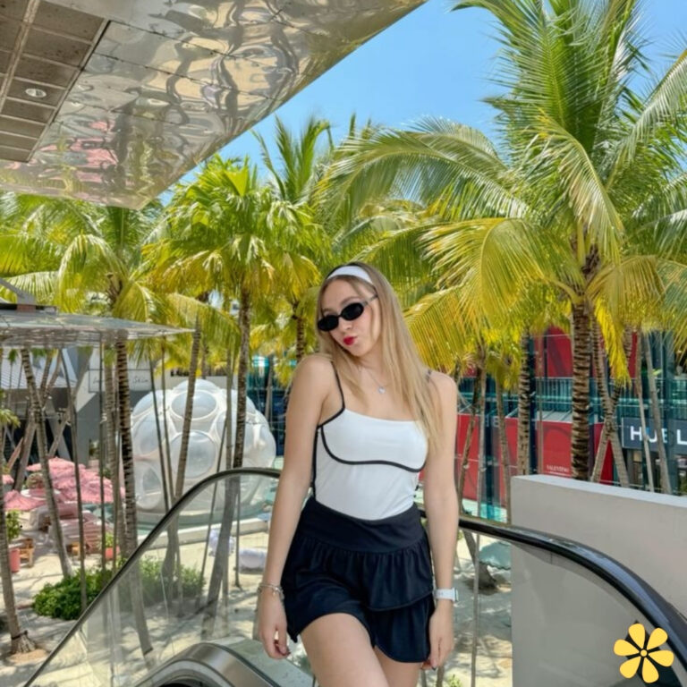 Woman in sunglasses and a white tank top poses playfully on an escalator surrounded by palm trees.