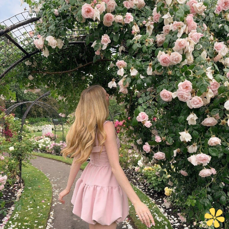 A woman in a pink dress walks under a blooming rose arch.