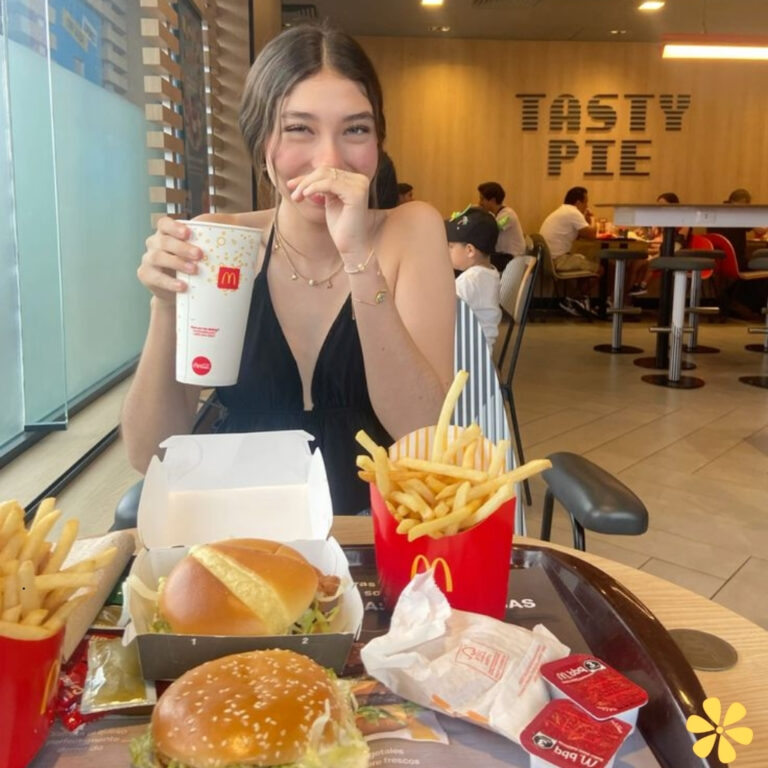 Smiling young woman enjoying fast food with fries, burger, and drink at a lively restaurant.