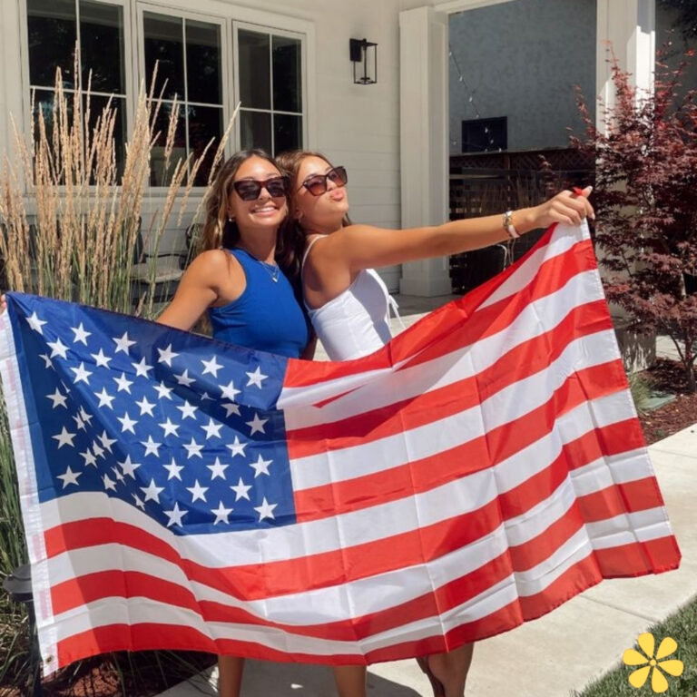 Two women holding an American flag, smiling outdoors with greenery in the background.