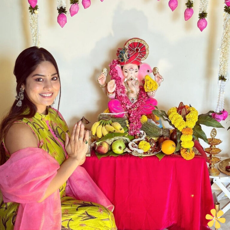 Woman in traditional attire prays before a colorful Ganesh idol surrounded by fruits and decorations.