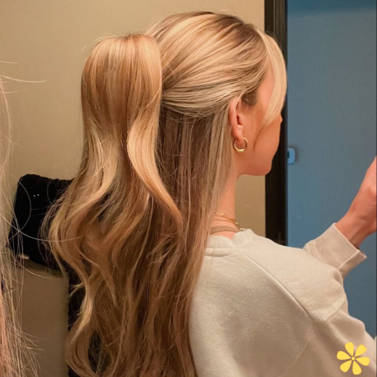 A woman with long, wavy blonde hair styled in a half-up ponytail stands in front of a mirror.