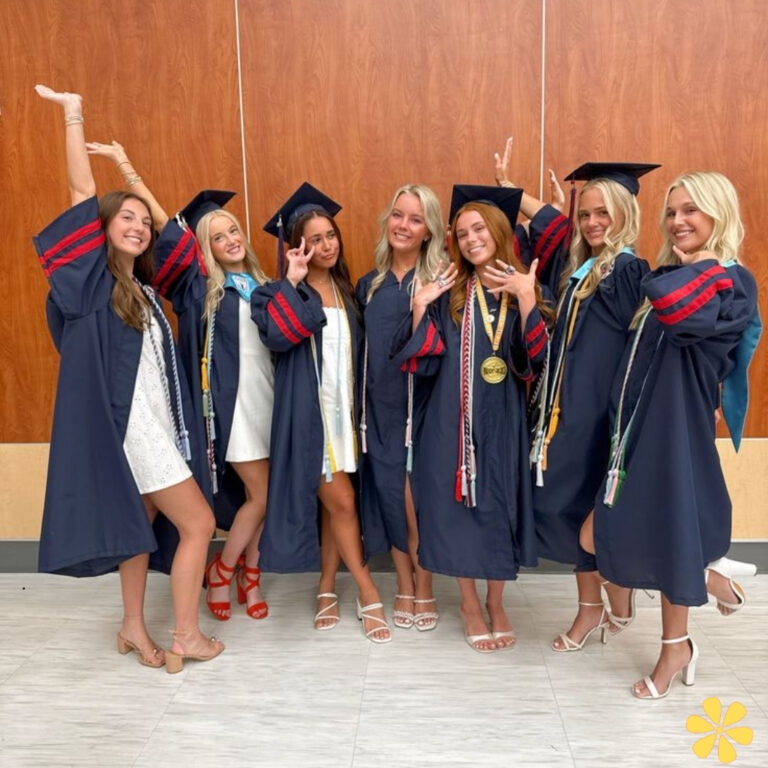 Seven graduates in navy gowns celebrate together, posing joyfully with hands raised.