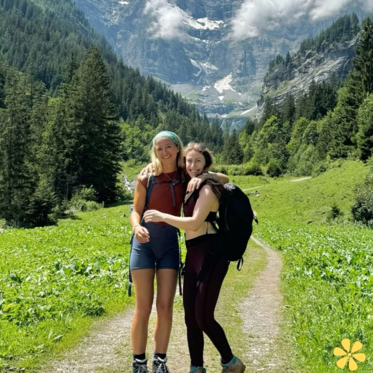 Two friends smile in a lush green valley, mountains towering behind them, enjoying a sunny hike.