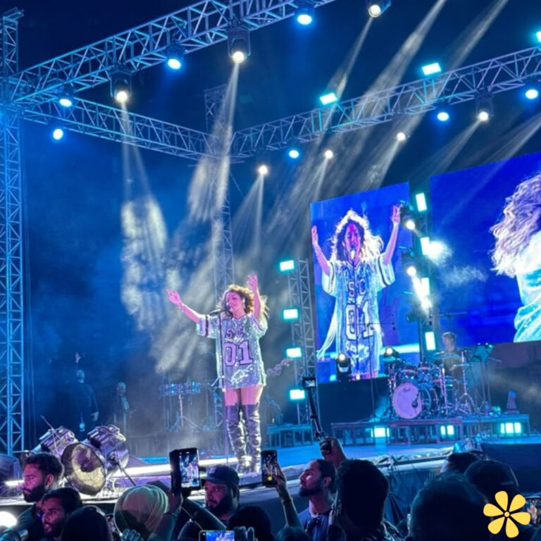 Performer on stage with arms raised, illuminated by bright lights and screens behind them.