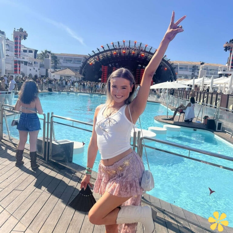 Young woman poses by a pool at a lively outdoor event, flashing a peace sign with a big smile.