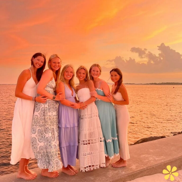 Six friends pose together by the sunset, each in beautiful dresses, smiles bright against the orange sky.