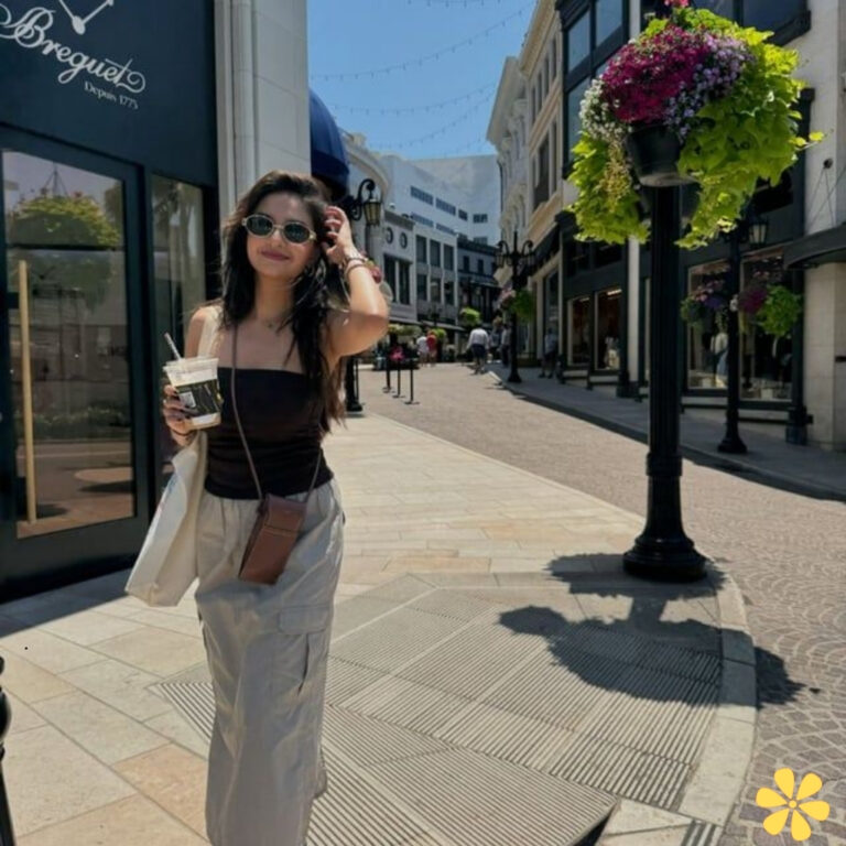 Young woman holding a drink, walking on a sunny street, shops on either side.