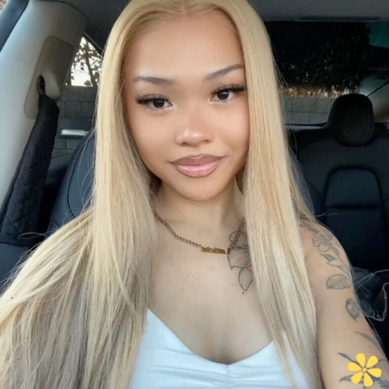 Smiling young woman with long, straight blonde hair, wearing a white top, seated in a car.