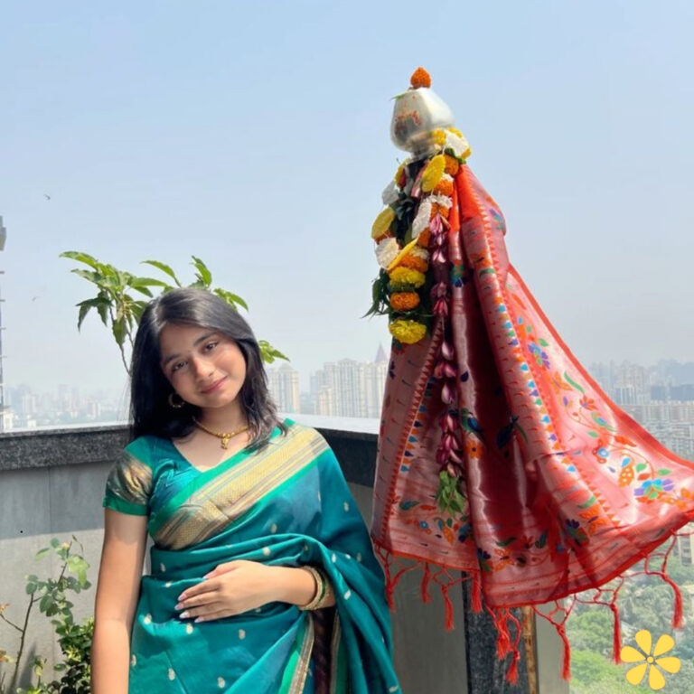 Young woman in a teal saree poses confidently, with a decorative pot and draped fabric beside her.